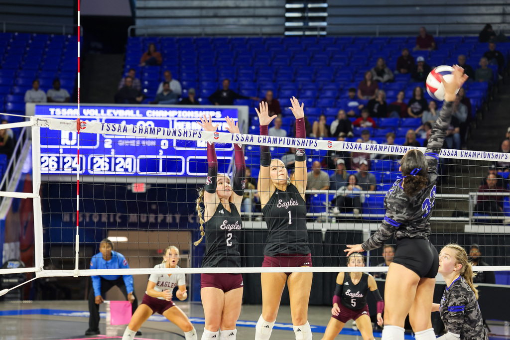 Several women playing volleyball. Two women jumping to block ball, one woman spiking. Players in maroon uniforms.