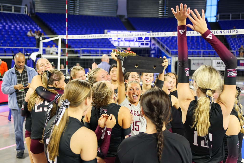 A team of women celebrating with a trophy, a man watching from the side, and a net in the background.