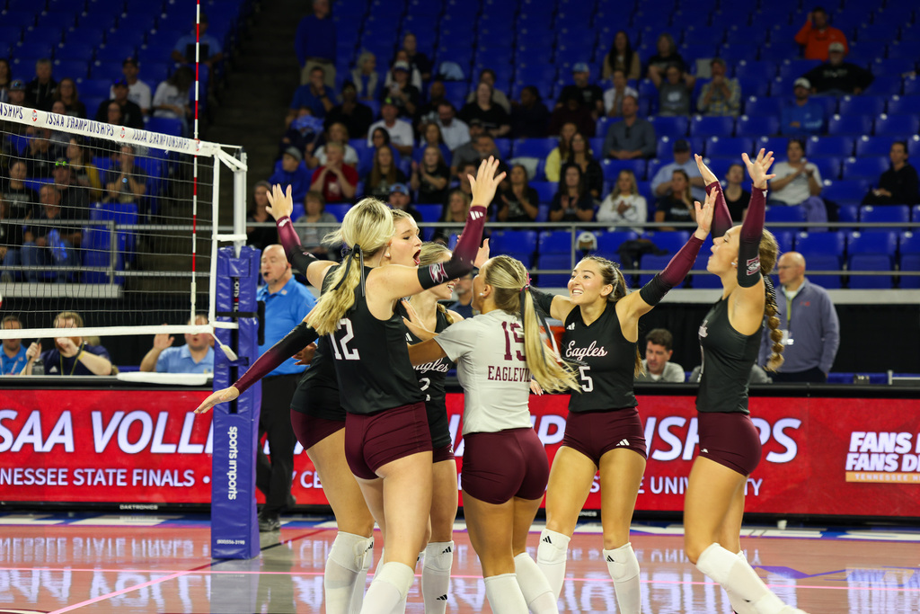 Volleyball team on a court celebrating; two with arms up, one hugging, two jumping. Audience in blue seats behind.