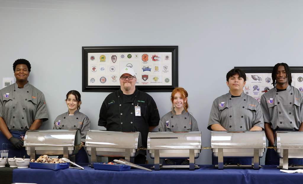 A group of people in gray uniforms stand behind a table with several chafing dishes.
