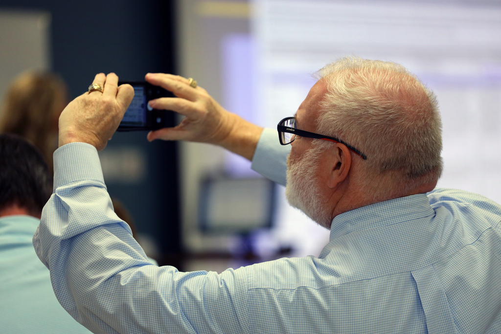 Man in blue shirt holding smartphone in both hands, looking at screen, standing in classroom.