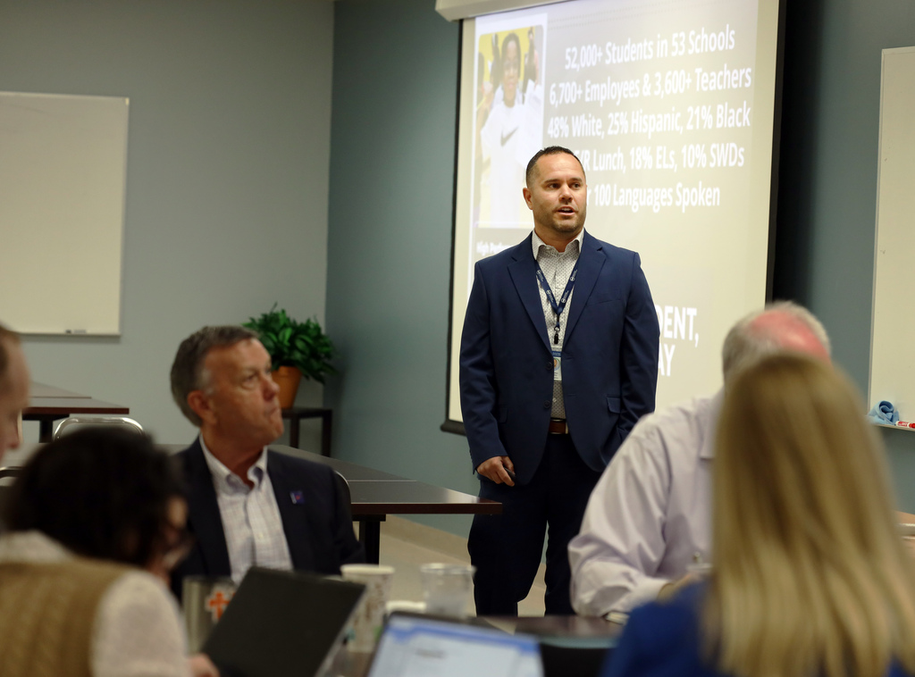 A group of adults in suits seated around a table with laptops; a man stands in front of them in a room with a presentation behind him