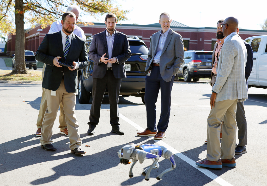 Six adult males in suits in parking lot, one with remote control for robot, others looking at robot