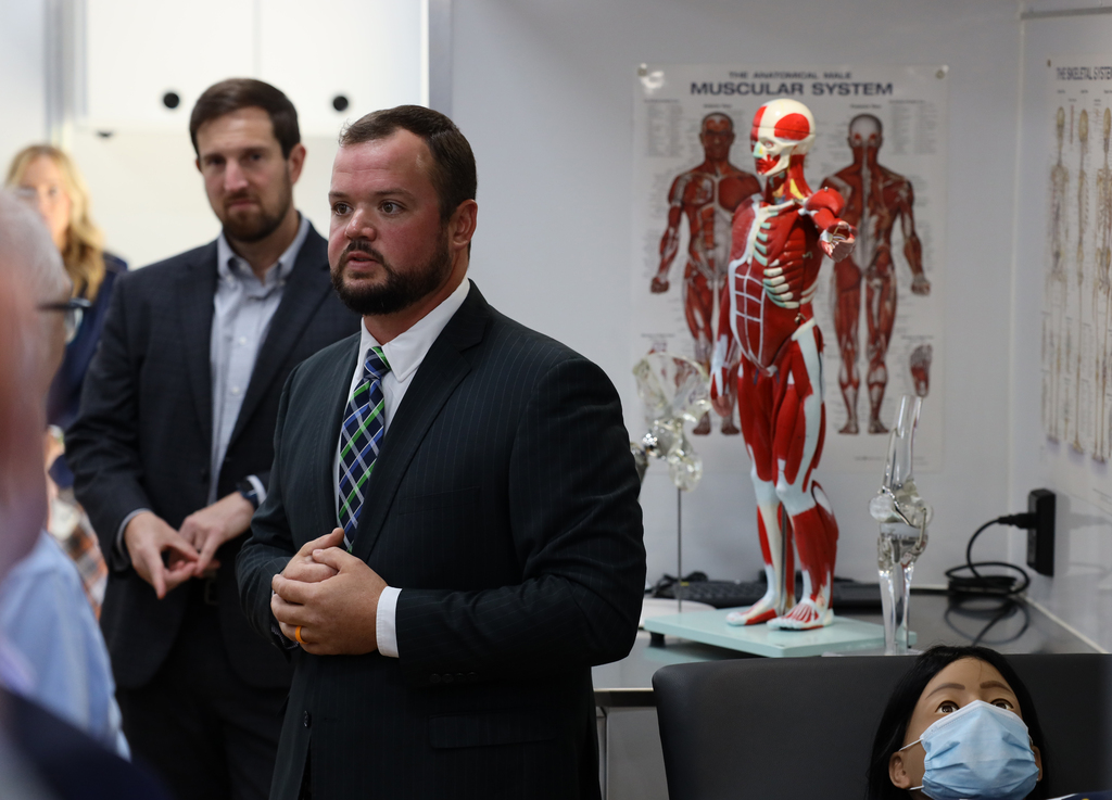 Three men in suits stand near a woman in a mask; one man gestures. Behind them, a model of a human skeleton is displayed on a table.