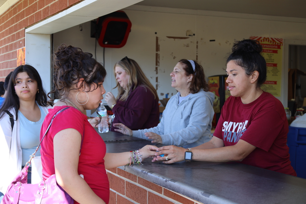 Five women at a counter, one giving a handshake. Behind them is a brick wall with a door.