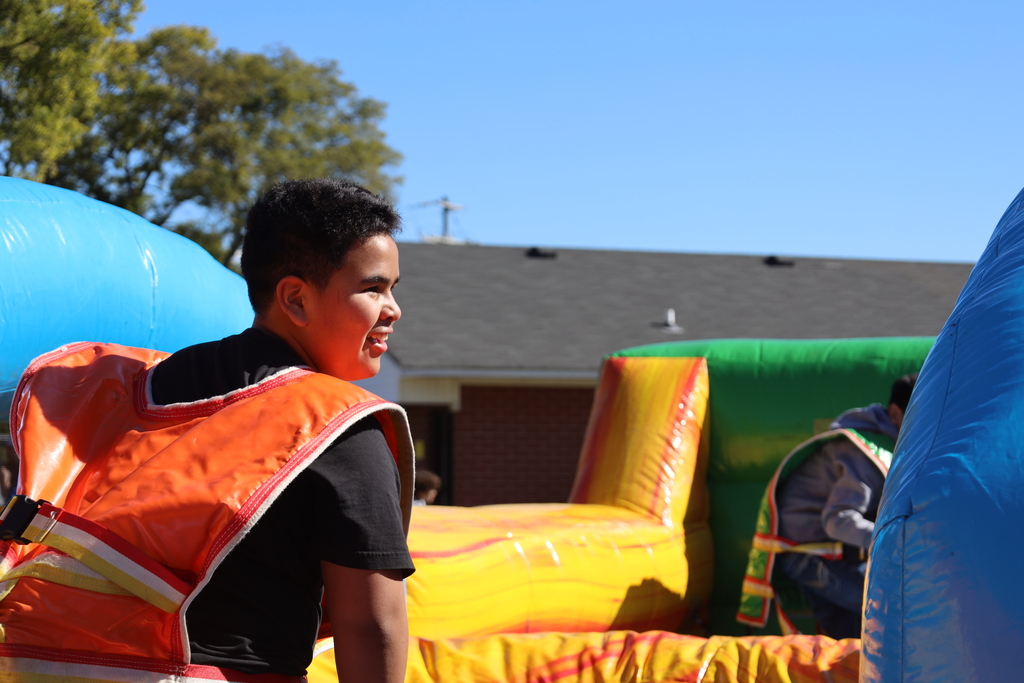 A boy in a safety vest looks at a bouncy house under a blue sky with trees.