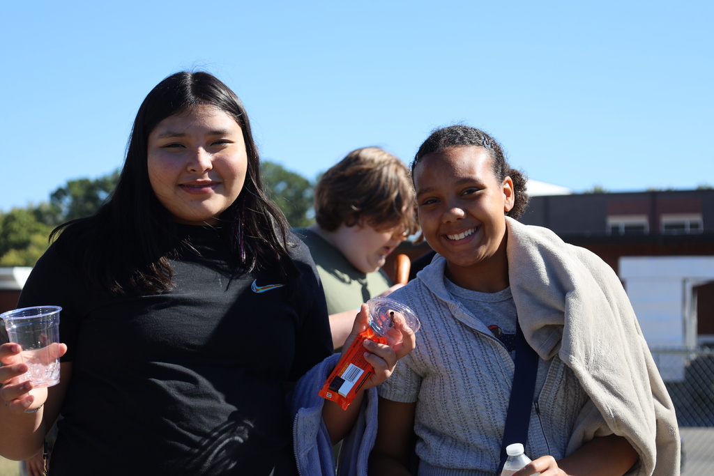 Three students outside on a sunny day, two of them holding drinks, with a fence and building in the background.