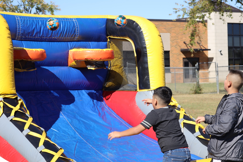 Two boys play on a blue, red, and yellow inflatable slide with basketball hoops; one slides, the other watches.