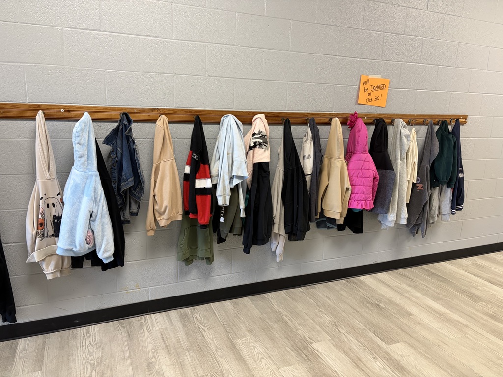 A row of various jackets and coats hangs on hooks against a white wall, on a wooden rod. A yellow sign is pinned to the wall.