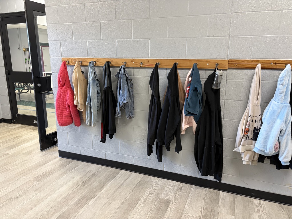 Inside a room, clothes are hanging on a wooden rack against a white wall. Floor is wooden.