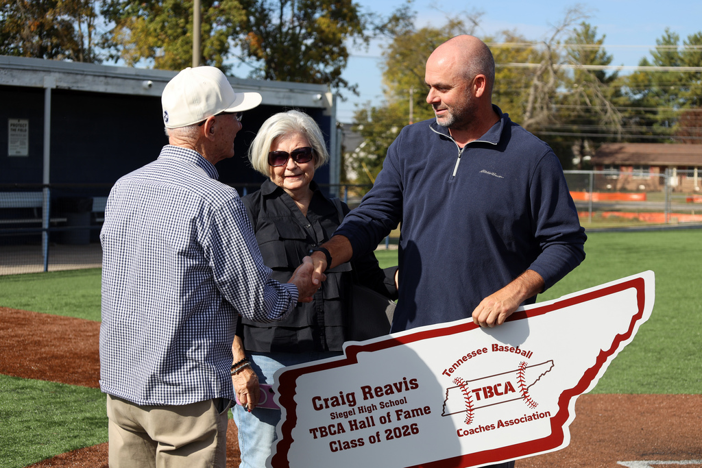 Three adults standing on a baseball field, one holding a sign, and shaking hands with each other.