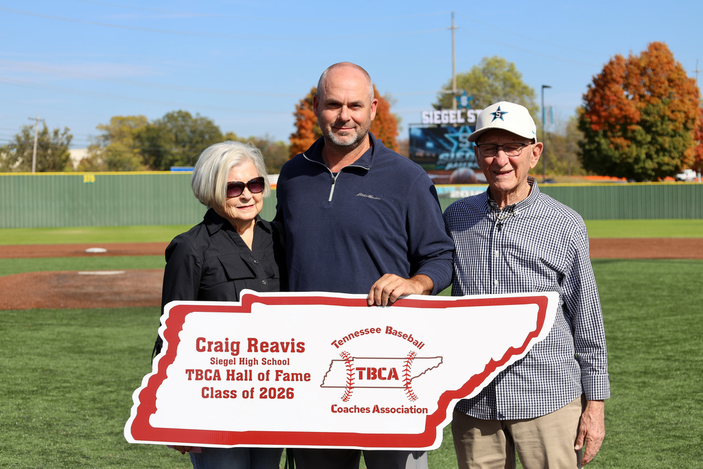 Three people pose with a plaque on a field, one person holding a sign. Green trees in background.