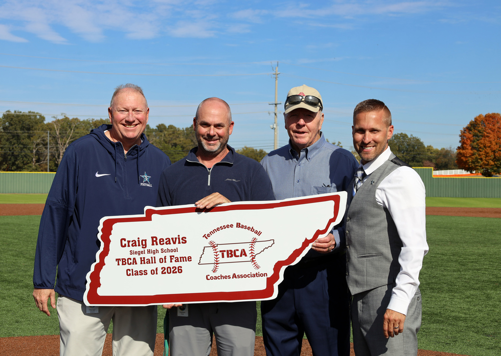 Four men stand on a baseball field, holding a large sign in front of them.