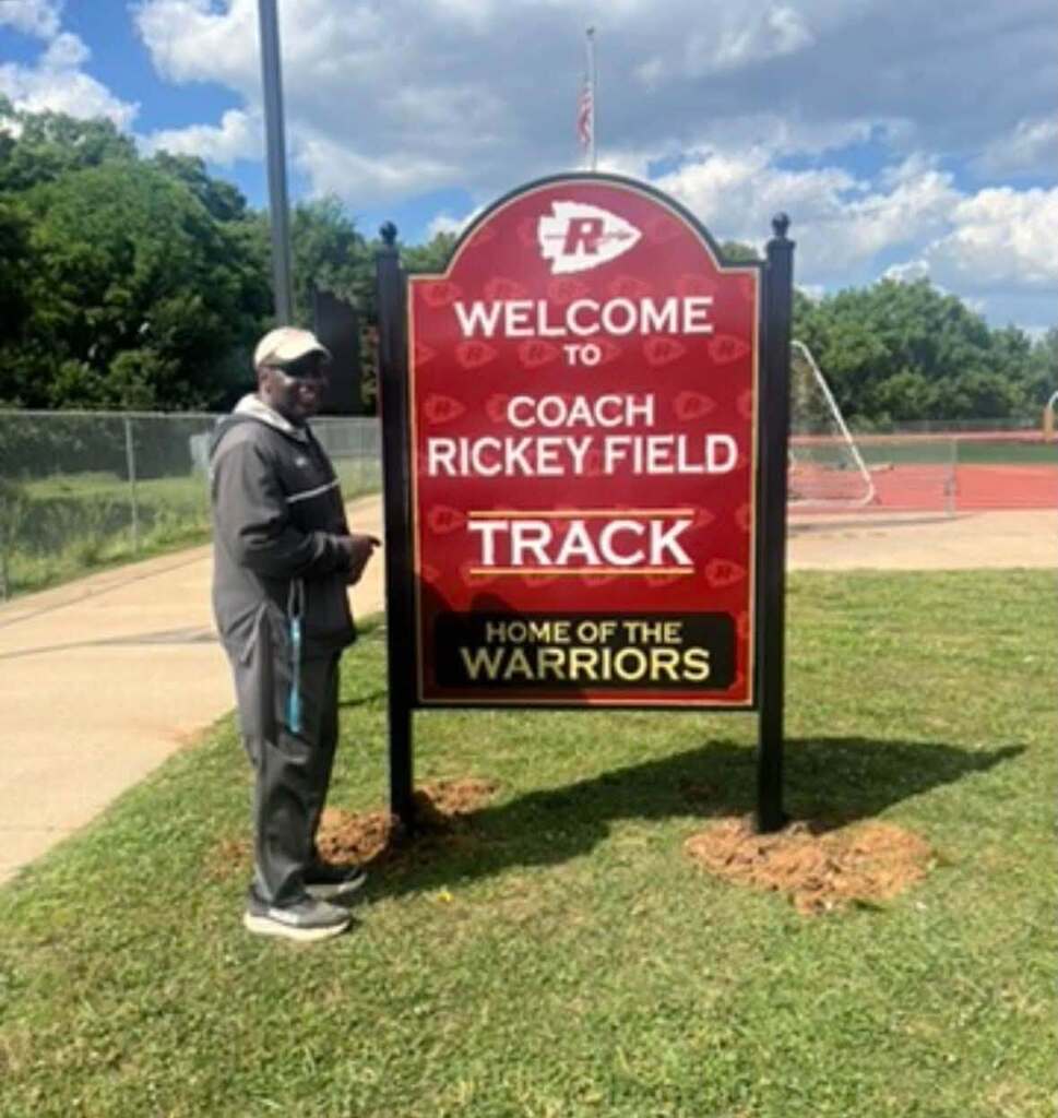 Rickey Field standing next to a sign that reads "Welcome to Coach Rickey Field Track". The sign includes the text "Home of the Warriors".