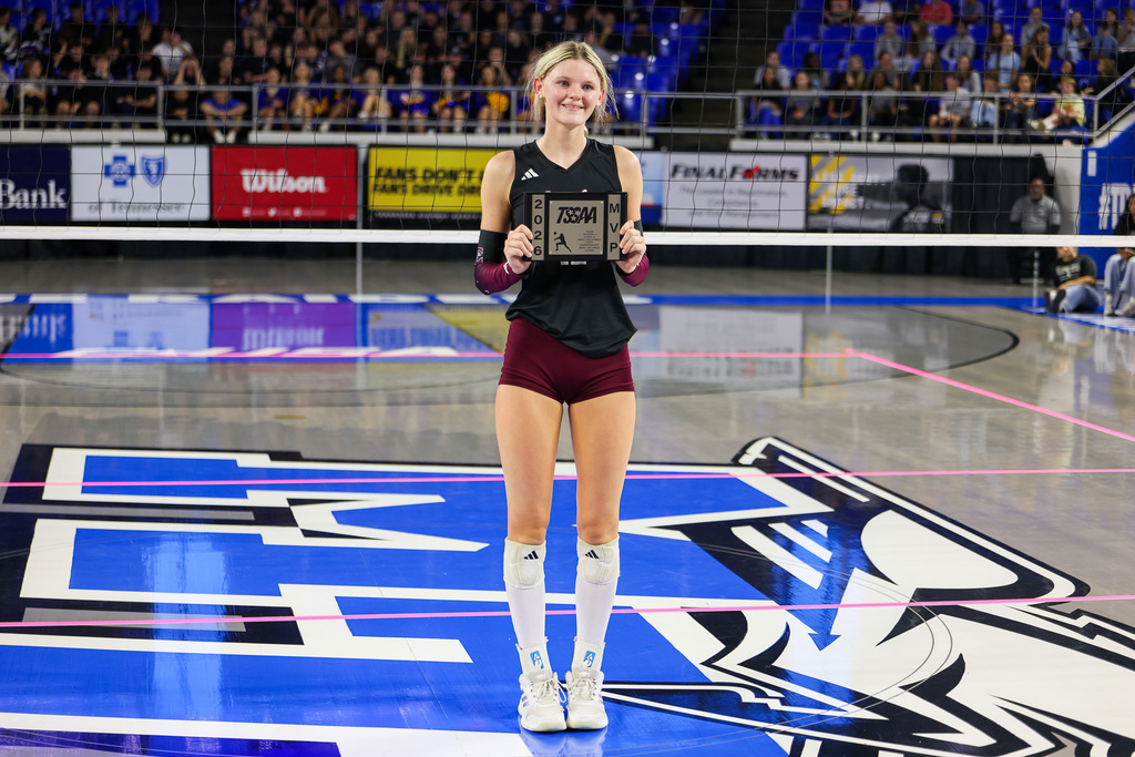 A female athlete stands in a sports court holding a framed award. She wears a black jersey, maroon shorts, and white socks.