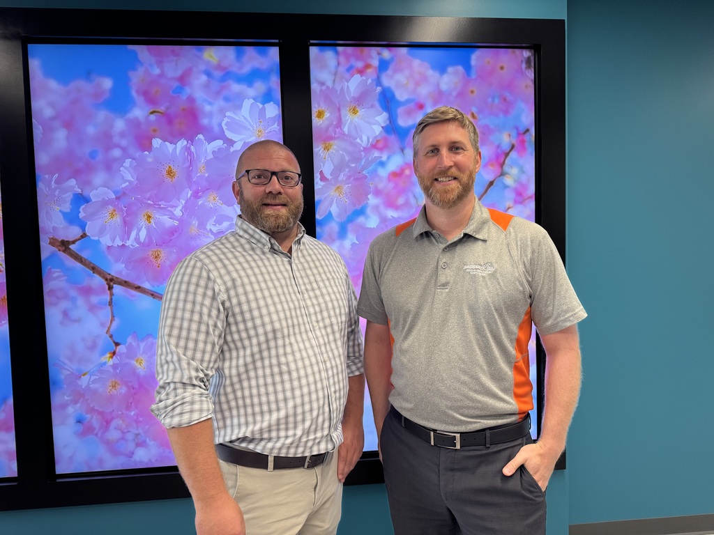 wo men stand side by side smiling in front of a wall featuring large panels with bright pink cherry blossoms against a blue sky. The man on the left wears glasses, a light gray and white checkered button-down shirt, and khaki pants. The man on the right wears a light gray polo shirt with orange accents and dark gray pants, with one hand in his pocket. The background wall is teal, creating a colorful and modern indoor setting.