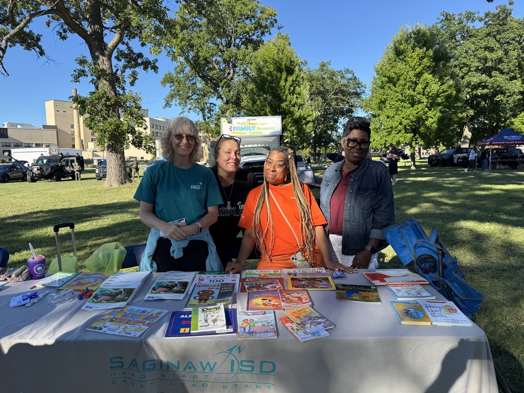 Four women stand behind a table outdoors at a community event, smiling at the camera. The table is covered with children’s books, flyers, and educational materials. The tablecloth reads “Saginaw ISD Head Start / Early Head Start.” In the background, people, tents, and vehicles are visible on the grassy park area with large trees and a clear blue sky.
