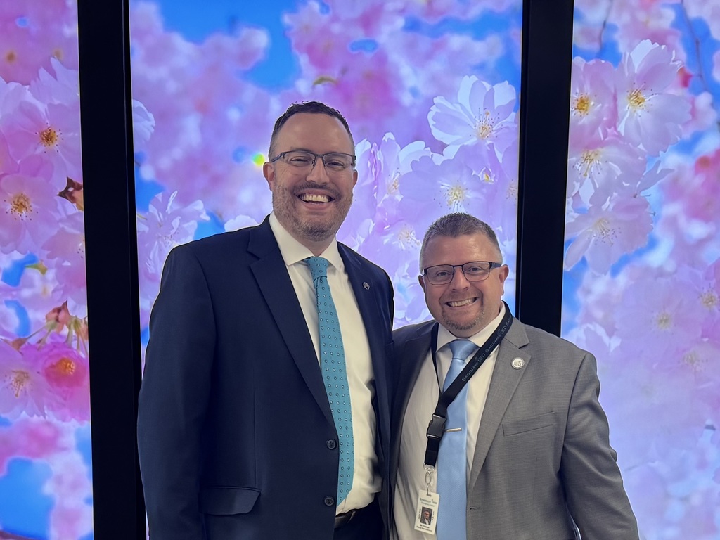 Two men in suits smiling and standing close together in front of a bright digital background filled with pink cherry blossoms. The taller man on the left wears a navy suit with a light blue patterned tie, while the shorter man on the right wears a gray suit with a solid light blue tie and a lanyard with an ID badge.