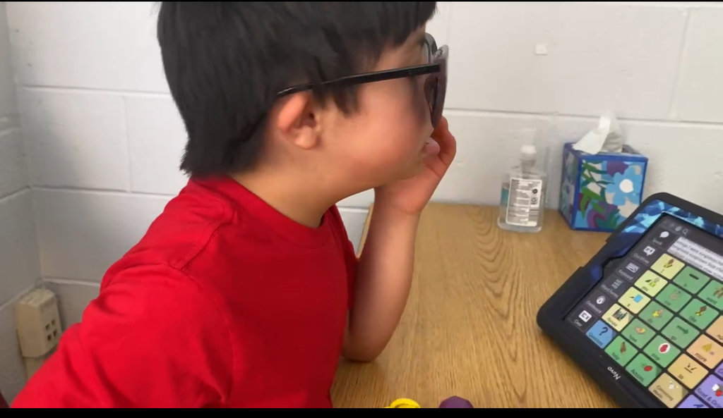 A young boy wearing glasses and a red shirt is sitting at a wooden table, touching his face while looking toward a tablet with an augmentative and alternative communication (AAC) app open. The table also has small colorful objects, a bottle of hand sanitizer, and a tissue box in the background.