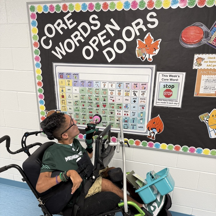 young adult male wheelchair user student presses a STOP button on a Core Word Bulletin board in a school hallway. 