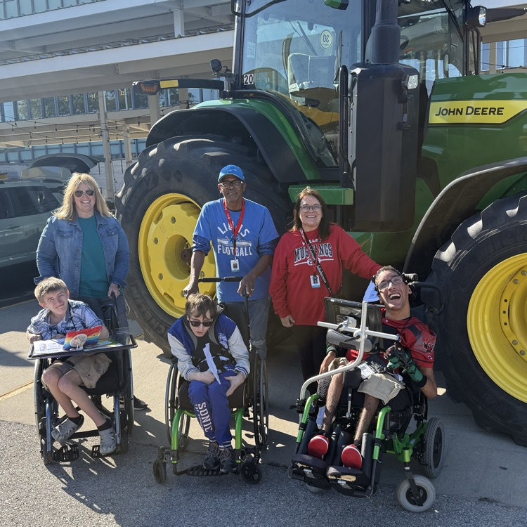 3 male wheelchair users with 2 female staff and 1 male staff posing in front of a large green and yellow tractor at an outdoor farmers market on a sunny day