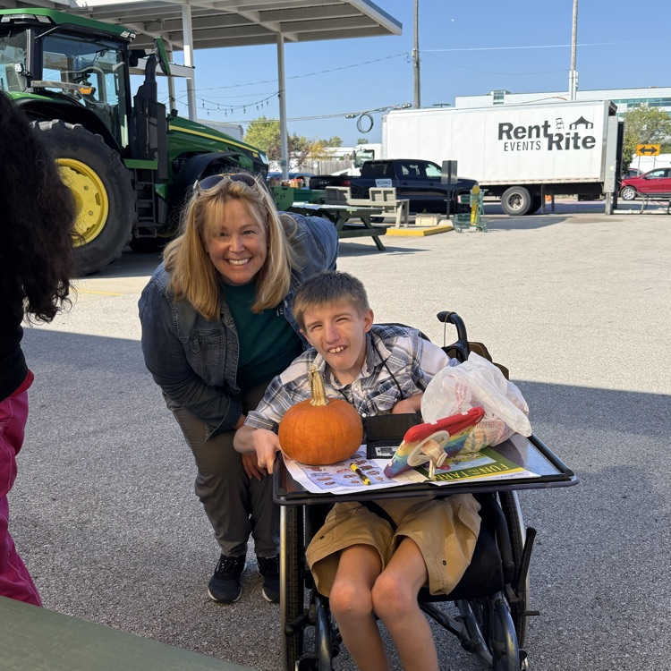 an adult male wheelchair user posing with his female teacher who is standing next to him. they are at an outdoor farmers market on a sunny day with pumpkins and other purchase from the market