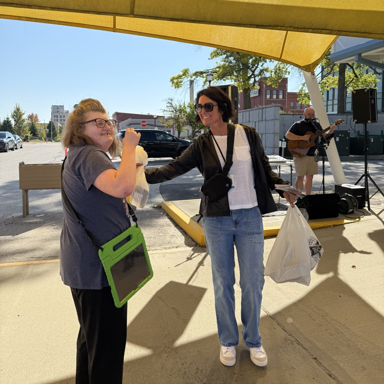 two adult females dancing to live music from a man with a guitar at an outdoor farmers market on a sunny day