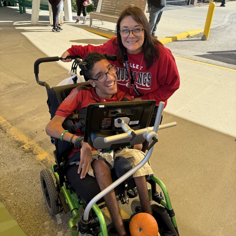 a male adult wheelchair user posing with his adult female teacher at an outdoor farmers market on a sunny day