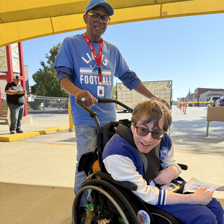 a male adult wheelchair user being pushed at an outdoor farmers market by a male staff on a sunny day