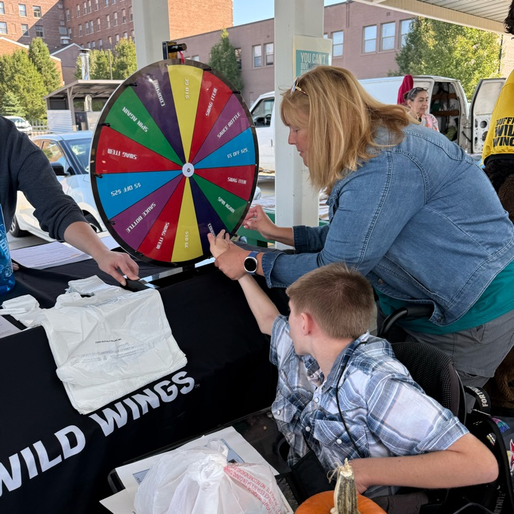 adult male wheelchair user getting help to spin a prize wheel at a booth at an outdoor farmers market on a sunny day