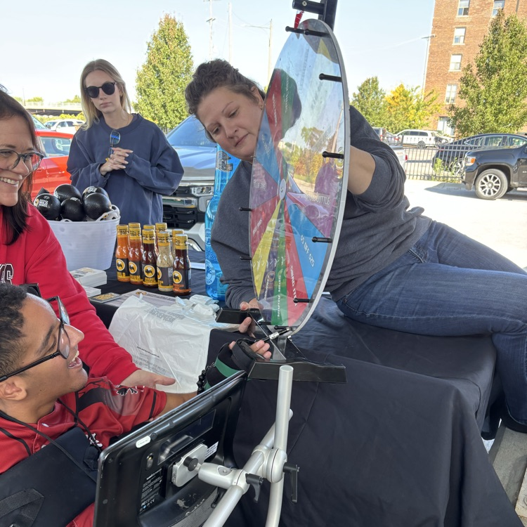 An adult male wheelchair user student with a female staff spinning a prize wheel at the outdoor farmers market