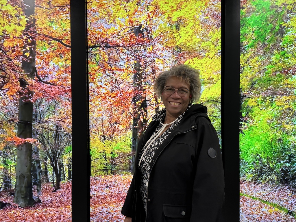 Fayerene “Faye” Meeks stands smiling in front of a vibrant autumn forest backdrop. The scene behind her features tall trees with golden, orange, red, and green leaves, creating a warm, colorful contrast to her black jacket and patterned shirt. She wears glasses and has short curly gray hair, radiating a cheerful and welcoming presence.
