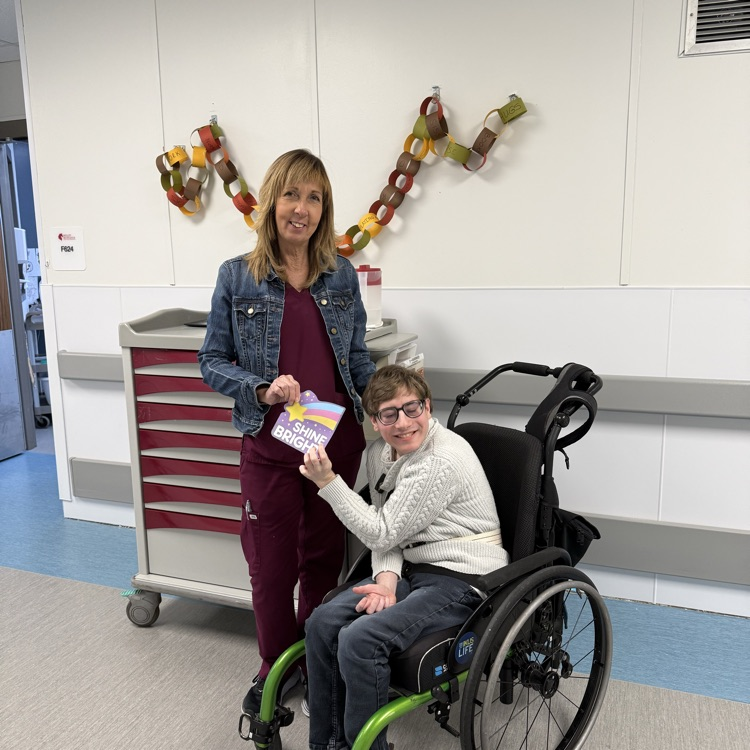 Young adult male student in a wheelchair hands a card to a female Nurse in a school hallway in front of a med cart  