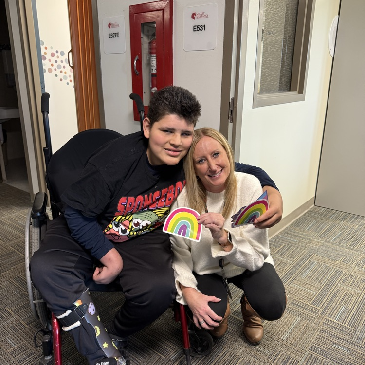 Adult female staff kneels down next to a young adult male student in a wheelchair. Staff is holding up the card she was given by the student who has his arm wrapped around her shoulder  