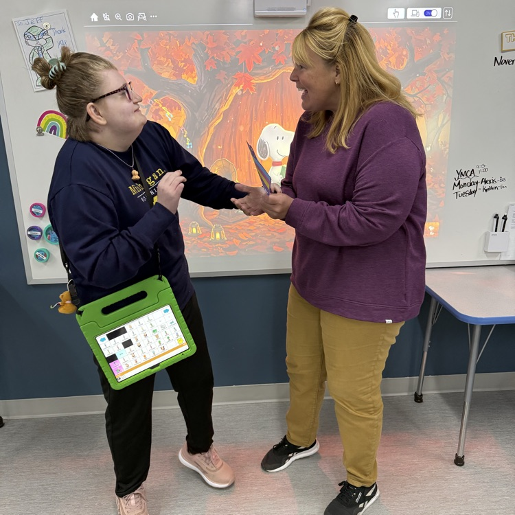 Young adult female student with a speech device hanging on her body hands a card to an excited female staff. The pair are standing in a classroom in front of a whiteboard with a fall scene. 