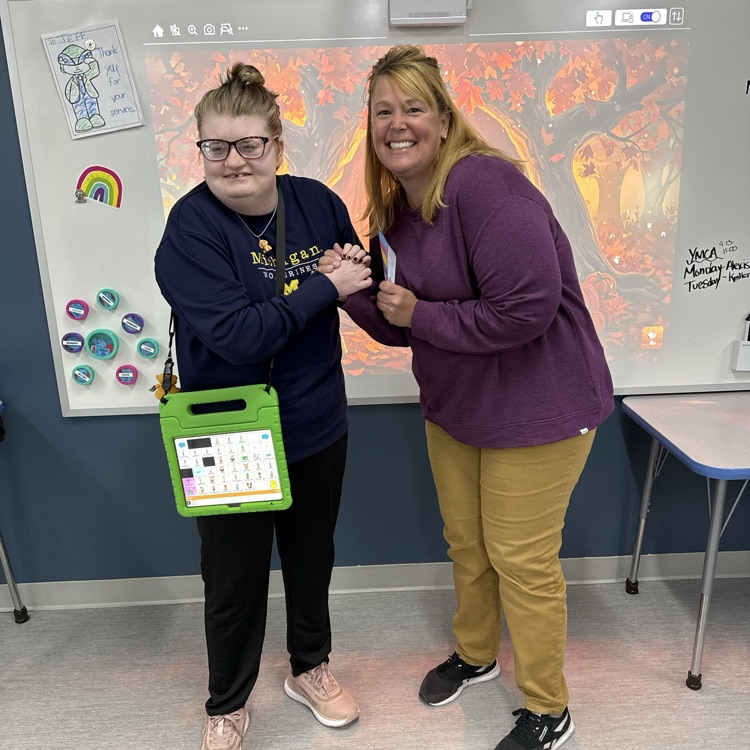 Young adult female student with a speech device hanging around her body poses with a female staff in front of a whiteboard in a classroom  