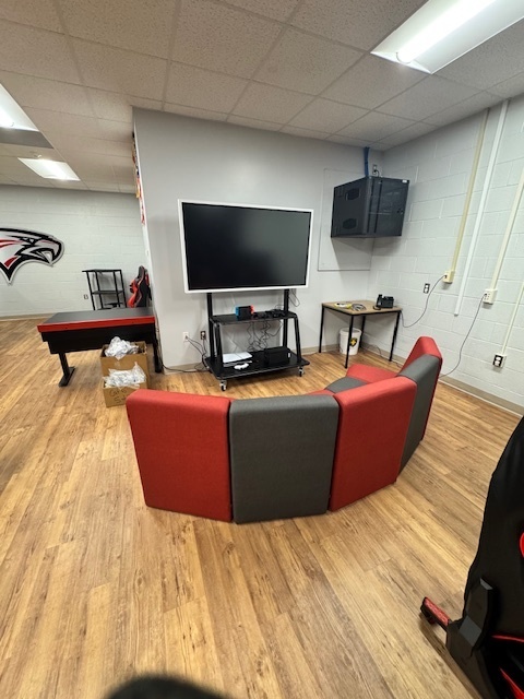 A photo of an esports lounge area featuring a large wall-mounted TV, a gaming console cart beneath it, and red-and-gray modular seating arranged in an L-shape. The room has wood-look flooring, white cinderblock walls, and additional tables and chairs along the sides.