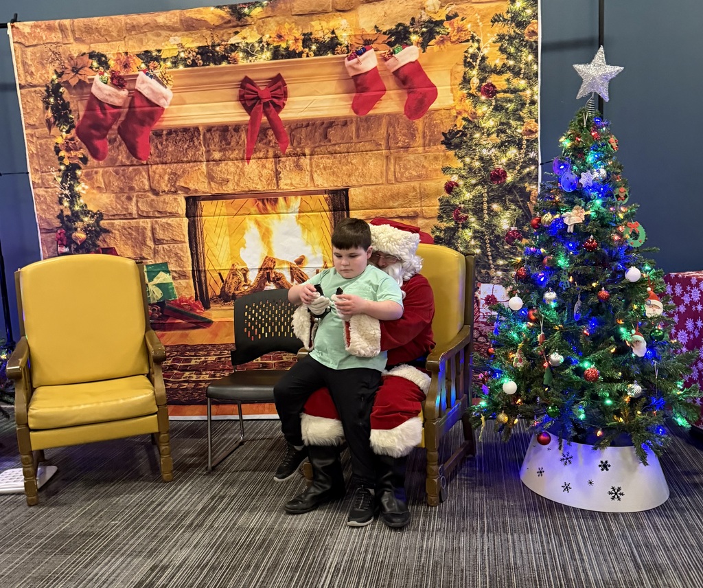 A child sits on Santa’s lap in a sensory-friendly holiday setup, holding a small object and looking down. Behind them is a printed backdrop of a cozy fireplace with stockings and holiday decorations. To the right is a small Christmas tree decorated with colorful lights and ornaments, and to the left are empty chairs, creating a calm, welcoming holiday scene.