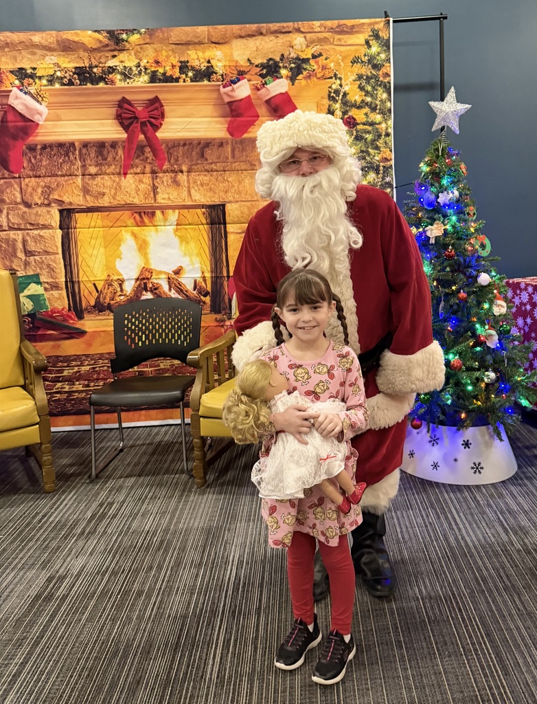 A smiling child stands in front of Santa, holding a doll dressed in a white outfit. Santa stands behind her with his hands gently at her sides. The scene is set in a sensory-friendly holiday space with a cozy fireplace backdrop, stockings and greenery above the mantel, yellow chairs nearby, and a small Christmas tree decorated with colorful lights and ornaments to the side.