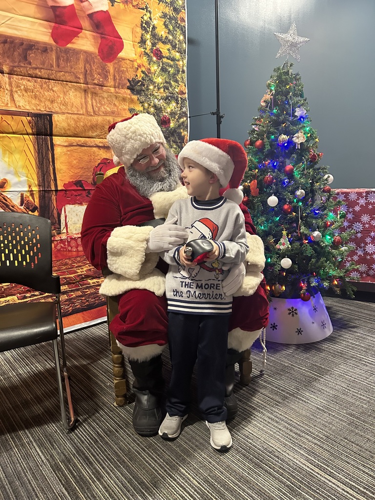 A child wearing a Santa hat stands in front of Santa, holding a small object while looking up at him. Santa sits behind the child, smiling and gently holding his hands. The scene is set in a sensory-friendly holiday space with a cozy fireplace backdrop, a decorated Christmas tree with colorful lights and a silver star topper, and chairs nearby, creating a calm and festive atmosphere.