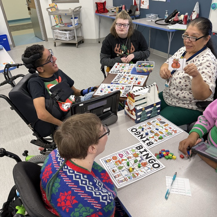 Adult wheelchair user students gathered around a table playing Holiday bingo in a classroom. Adult female staff is showing the next call picture
