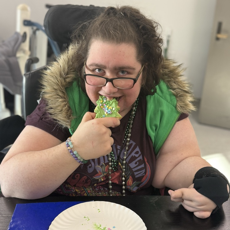 adult female wheelchair user wearing glasses and looking at the camera while eating a green frosted Christmas cookie