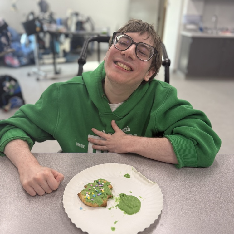 Adult male wheelchair user student in a green sweatshirt wearing glasses smiling at the camera with a green Christmas tree cookie in a plate in front of him