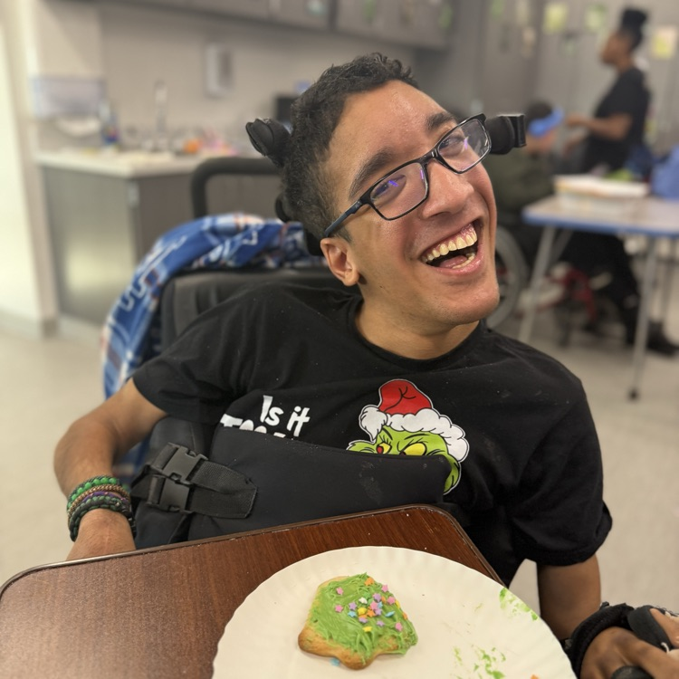 adult male wheelchair user student wearing glasses smiles at the camera with a plate in front of him that holds a green frosted Christmas tree cookie with sprinkles