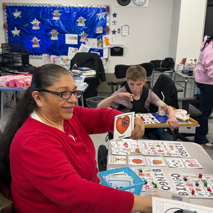 female parapro holding up a heart picture bingo card in a special needs classroom full of adult students with special needs