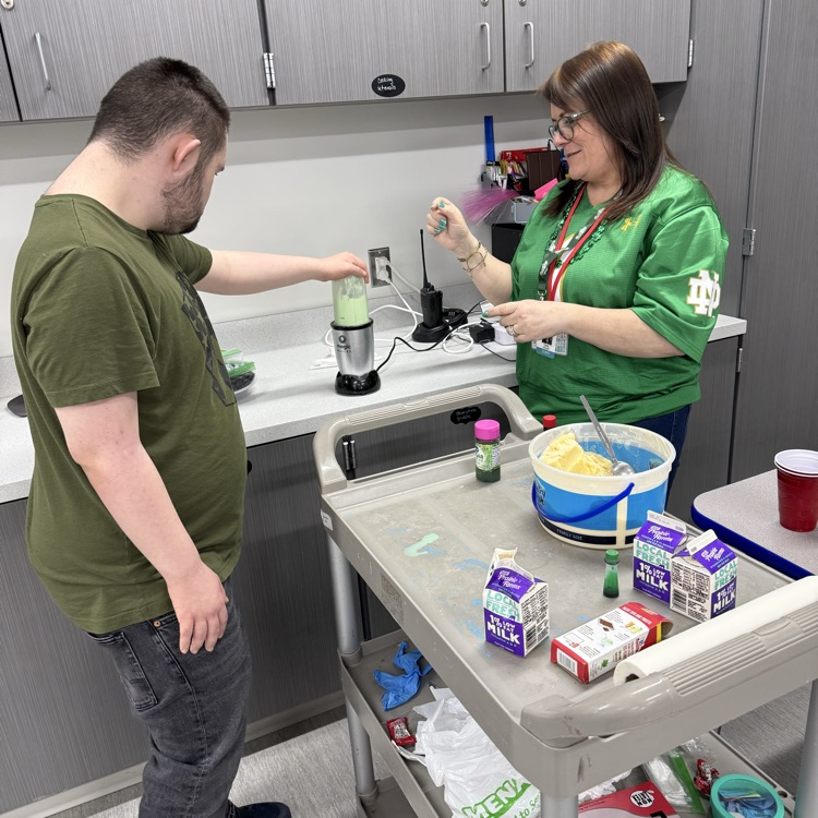 Adult male transition student uses a blender to make a Shamrock Shake. His teacher is helping and between them sits a cart with the ingredients  