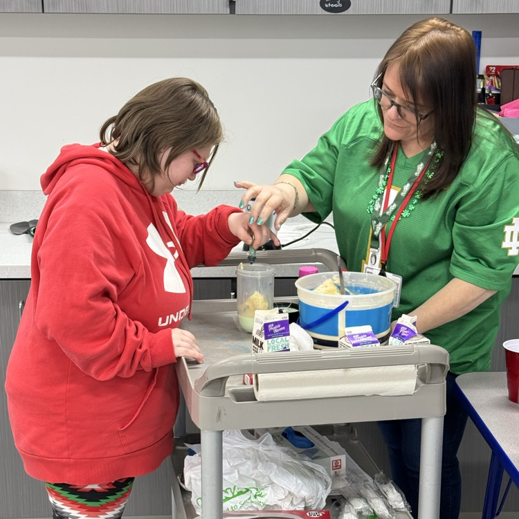 an adult female transition student adds green food coloring to her blender but to create a shamrock shake with her teacher’s help. they are assembling the shake on a cart sitting between them  