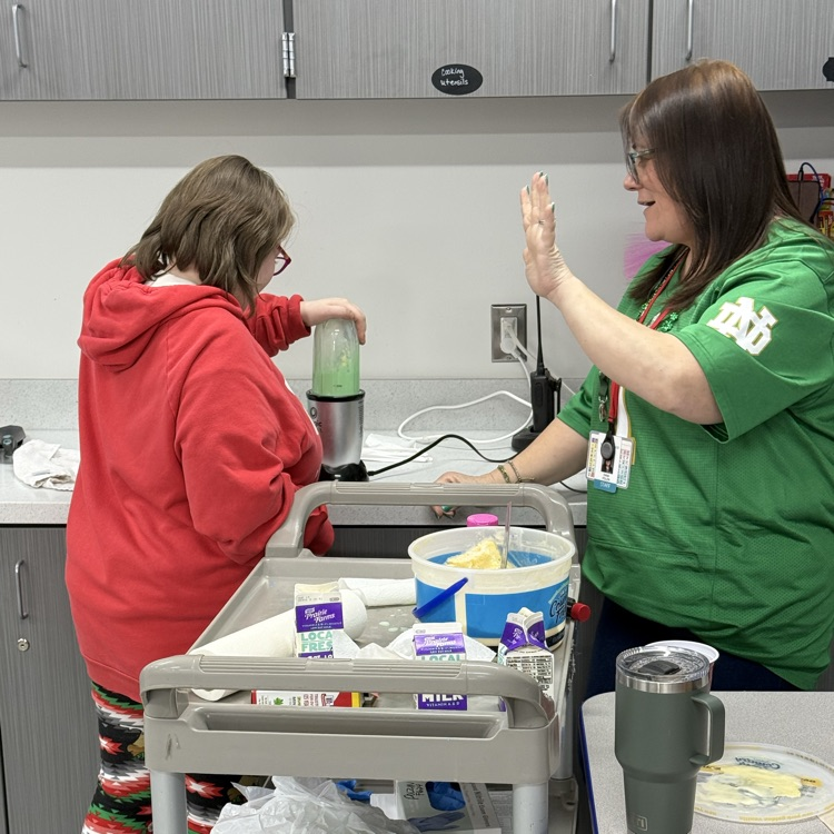 the female teacher counts out loud on her fingers how may more seconds of blending the adult female transition student needs to complete the recipe. the student is using a blender to make the shamrock shake. a cart sits between the two  
