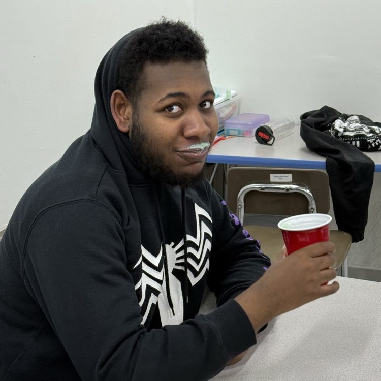 an adult male transition student sits at his desk smiling at the camera with a green shake mustache to show that he enjoyed the treat  
