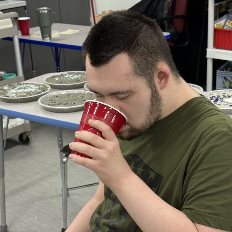 an adult male transition student drinks a shamrock shake from a red plastic cup while sitting at his desk in the classroom  